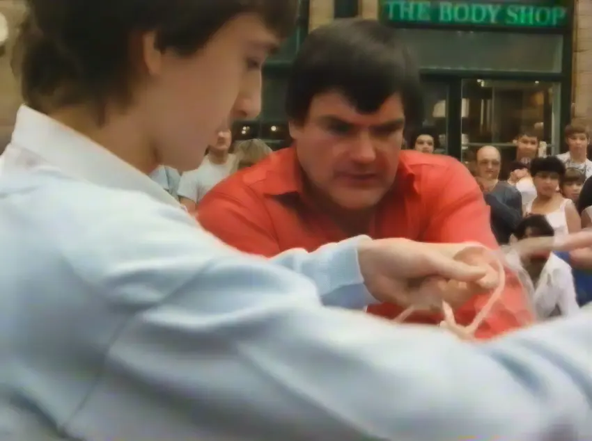 David Portsmouth (on left) and Doug Alker (centre) are shown with David tying Doug's hand up for a magic trick. The location is Covent Garden. The background has a number of onlookers interested to see what happens. Many do not even know both these guys are deaf!