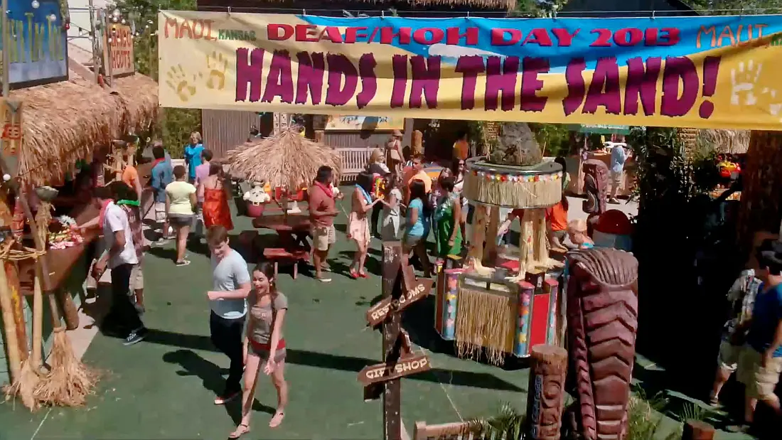 Fictional theme park Maui Kansas is depicted in this scene with banners and stalls. A large banner above the scene declares 'Deaf/HOH Day 2013' and 'Hands in the Sand!' Daniel Durrant can be seen at bottom left talking to oe of the Carlton school crew.